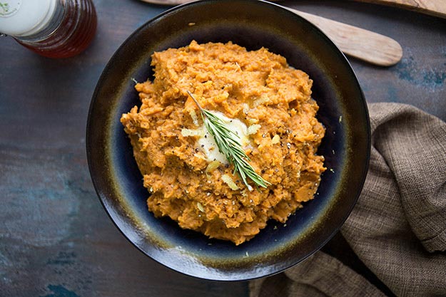 A bowl of mashed sweet potatoes with rosemary on a wooden table.