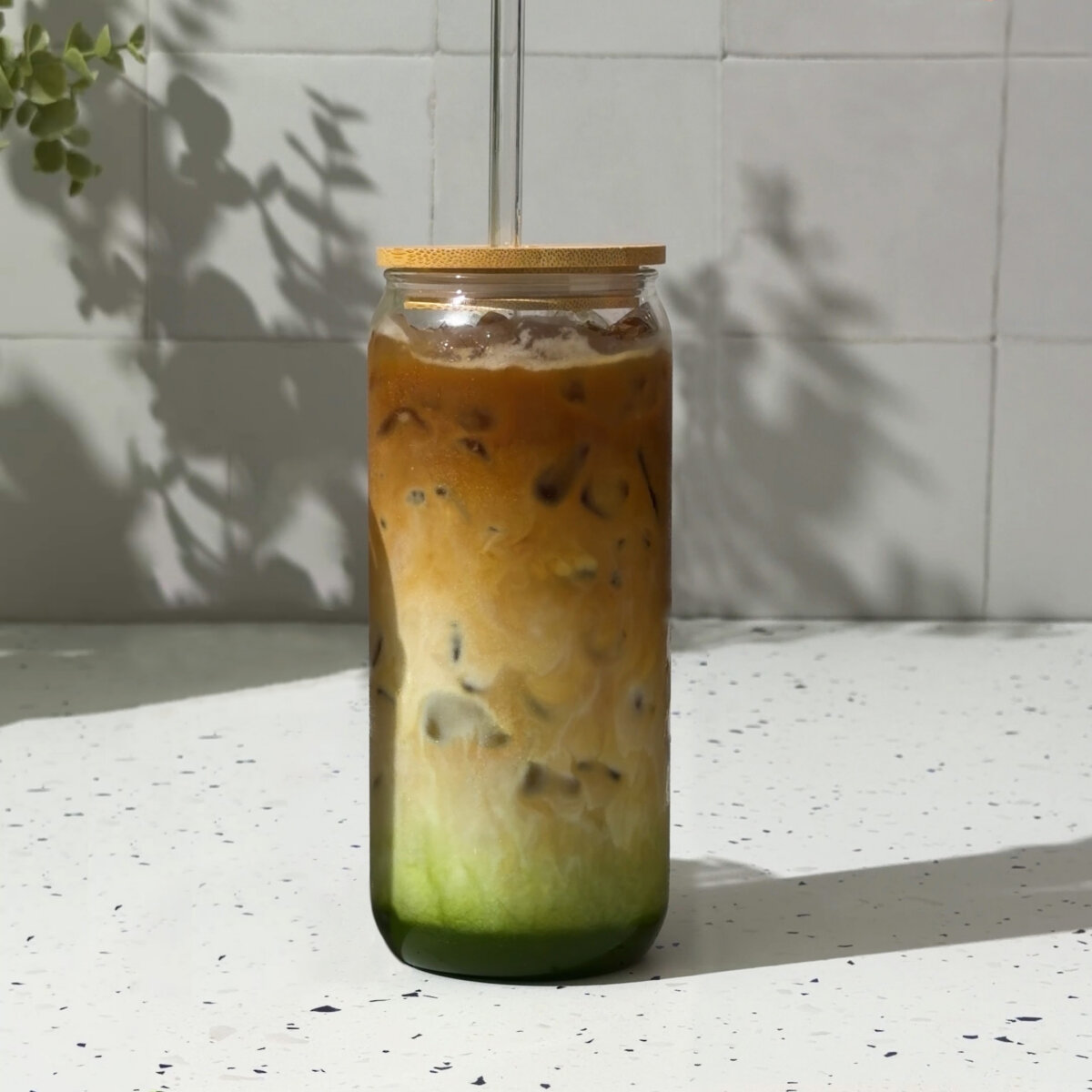 Glass of iced coffee with milk and espresso and matcha layers, shown as a hand gently lifts the glass on a bright kitchen counter.