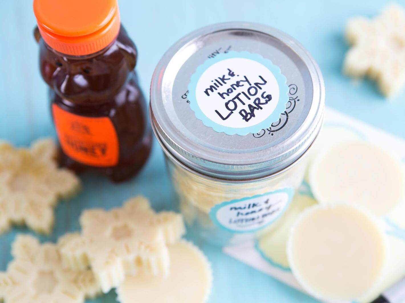 Homemade milk and honey lotion bars in a jar and scattered on blue surface, beside a bottle of Nate's honey.