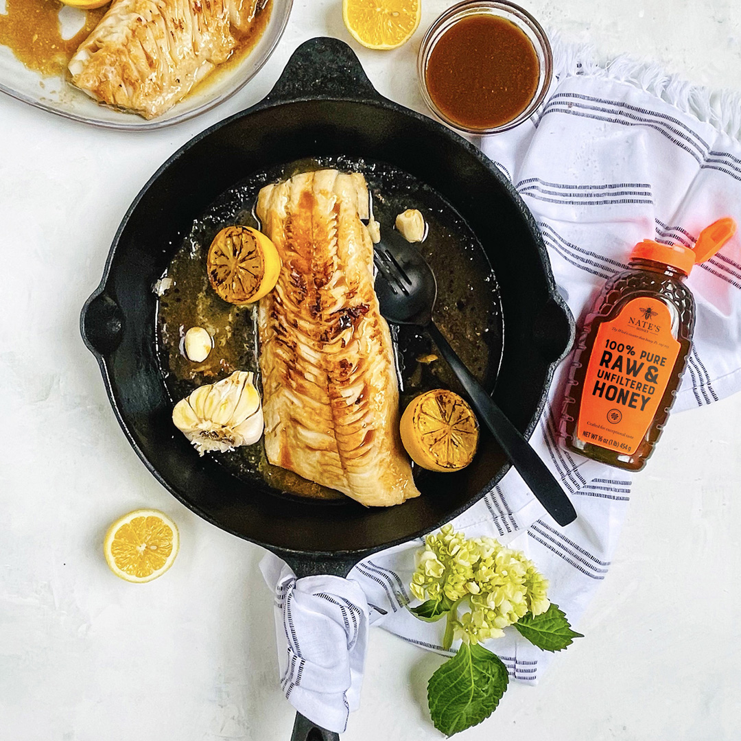 Pan-seared fish fillet in a skillet with lemon slices and garlic cloves, with bottle of Nate's Honey and a sauce bowl.
