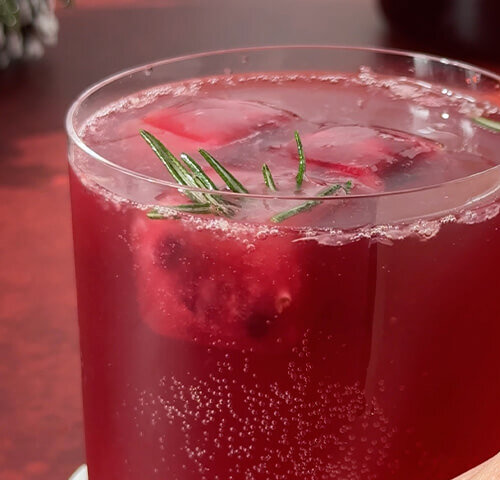 Cup filled with red Mistletoe Margarita Punch and rosemary, cranberry ice cubes.
