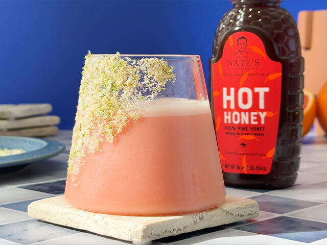 Pink drink in glass decorated with citrus salt garnish. Next to Nate's hot honey.