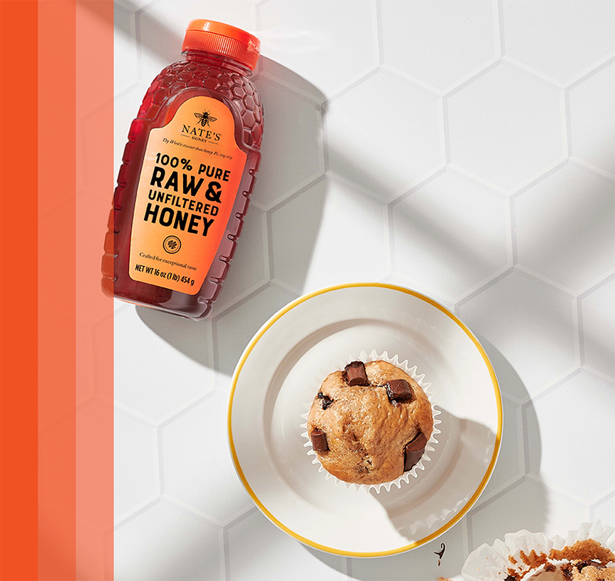 Nate's honey bottle beside a chocolate chip muffin on white hexagonal tiles.