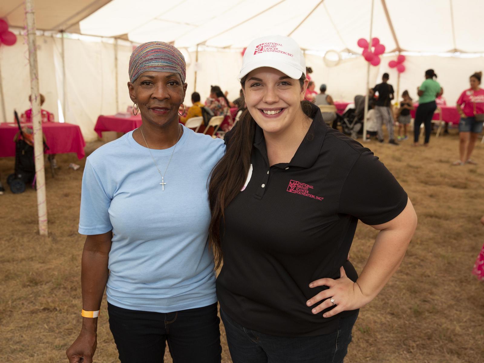 Two smiling women, one in blue and one in black with event branding, at an outdoor tent gathering.