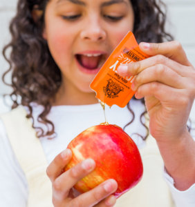 Child pours honey from a Nate's honey mini on apple, delight on face, vibrant colors focus.