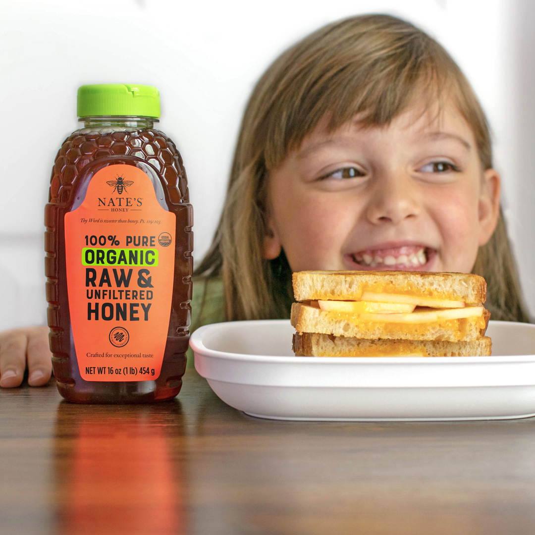 A smiling girl peeking behind a apple, cheese and honey sandwich beside a bottle of organic Nate’s Honey on a table.