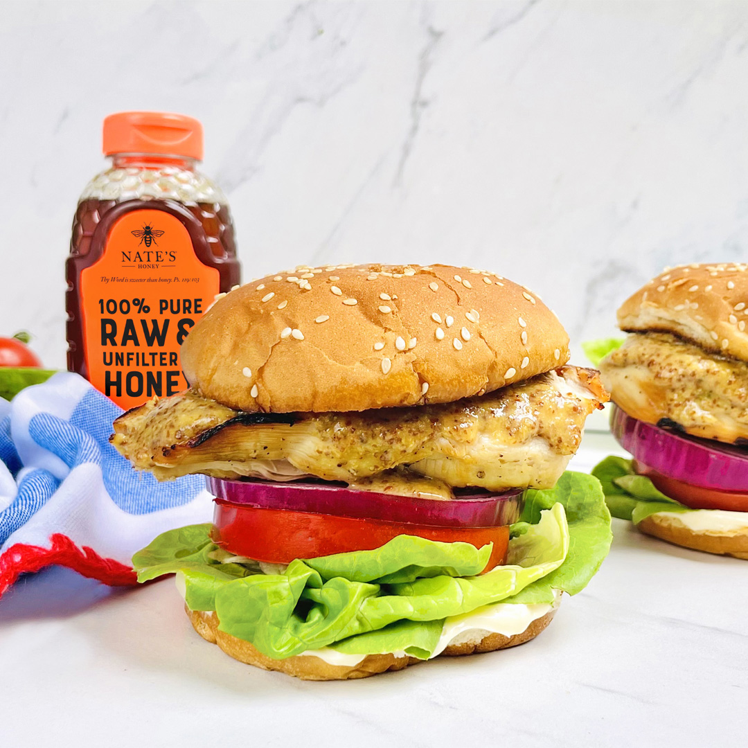 Hot honey mustard chicken sandwiches with lettuce and tomato, and a bottle of Nate’s Honey on a marble surface.
