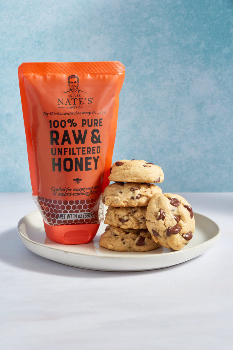 A pouch of honey beside a stack of chocolate chip cookies on a plate, suggesting the use of honey in the cookies.