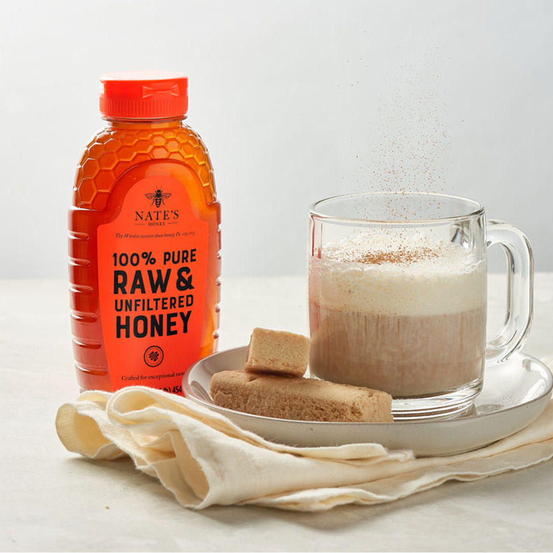 A bottle of Nate’s Honey next to a steaming mug of latte accompanied by shortbread cookies on a white background.