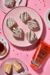 White cakes with chocolate drizzle on pink plates, Nate's honey pouch, and a cup of tea on a pink surface.