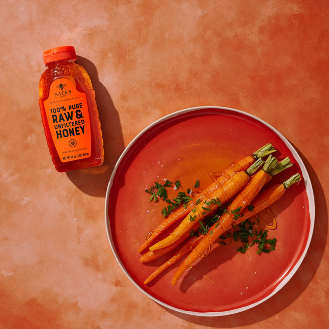 Honey-glazed carrots on a plate with Nate's Honey bottle on the side on an orange background.
