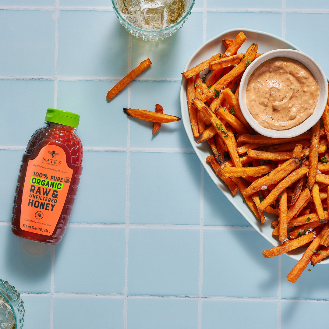 Sweet potato fries with dip and a bottle of organic Nate’s Honey on blue tiled surface, drinks in background.