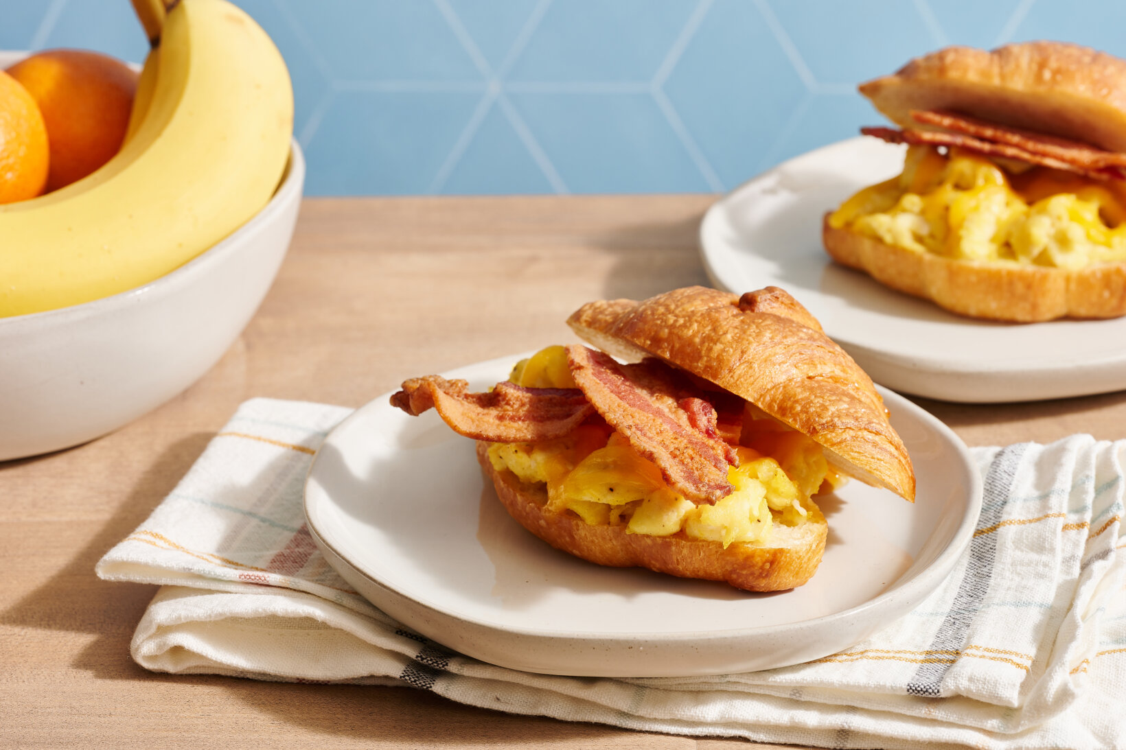 Egg and bacon croissant sandwiches made with honey on plates, a bowl of bananas, oranges on a wooden kitchen counter.