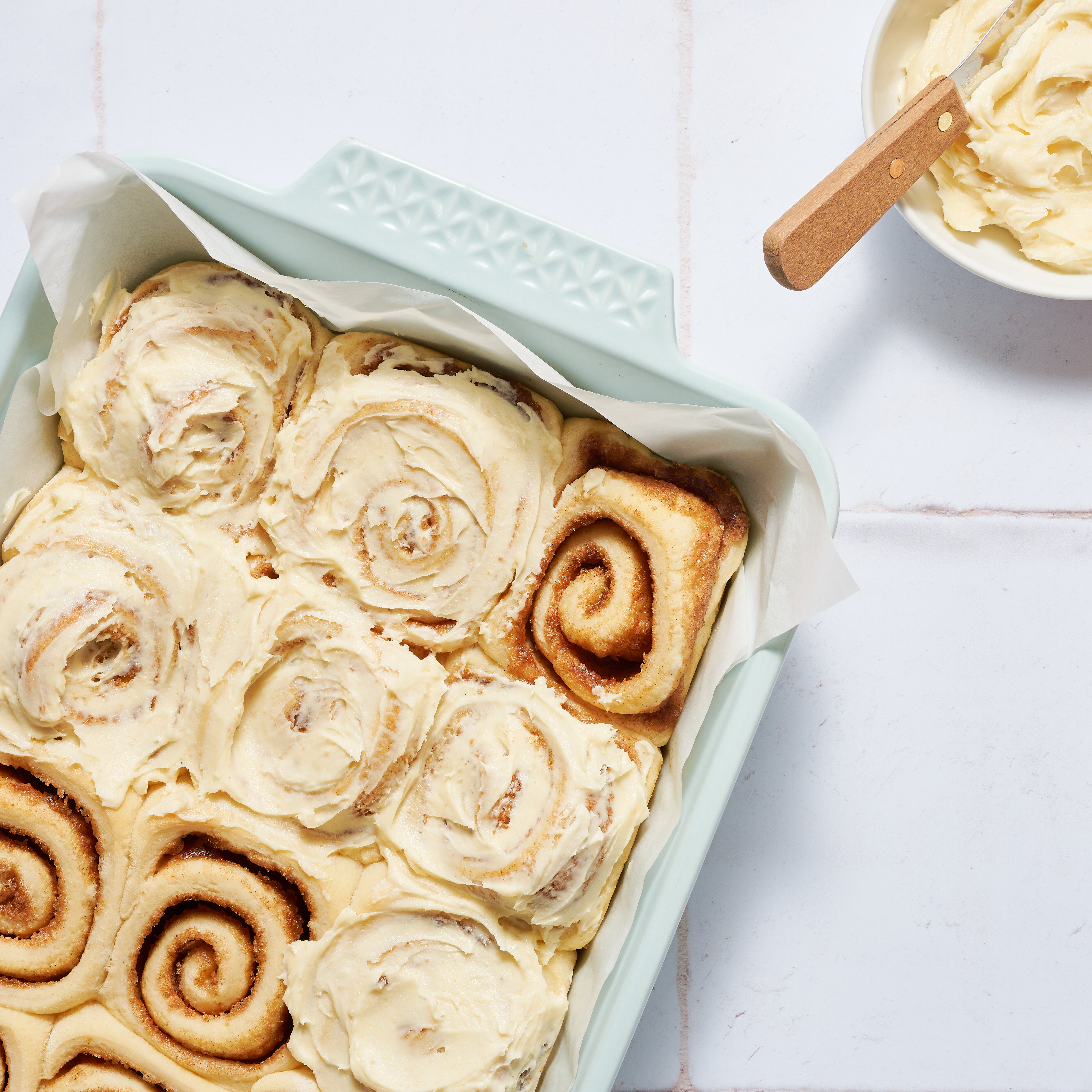 Honey cinnamon rolls with cream frosting in a baking dish, one roll unfrosted, on a white countertop.