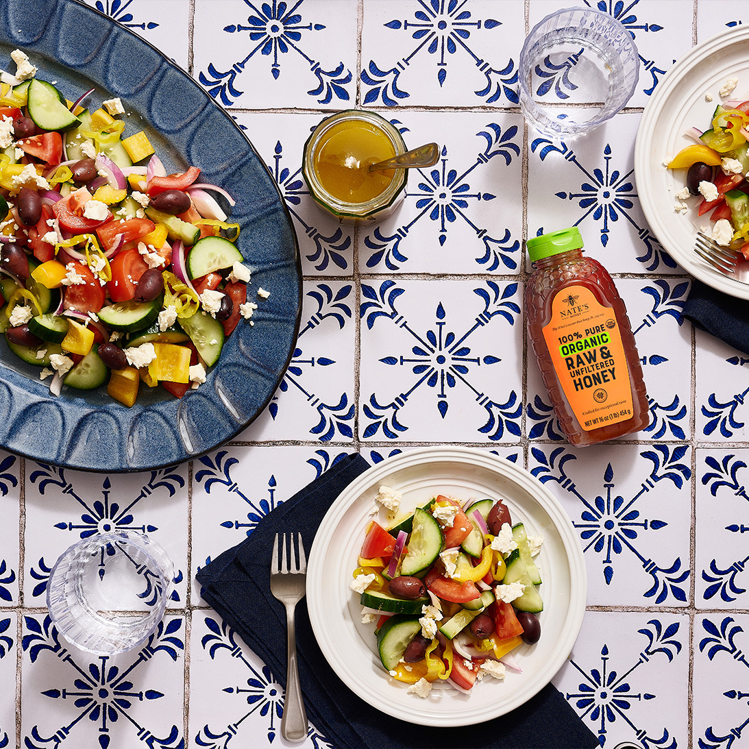 Greek salads, honey salad dressing and a bottle of Nate’s organic honey on a blue and white tiled table.