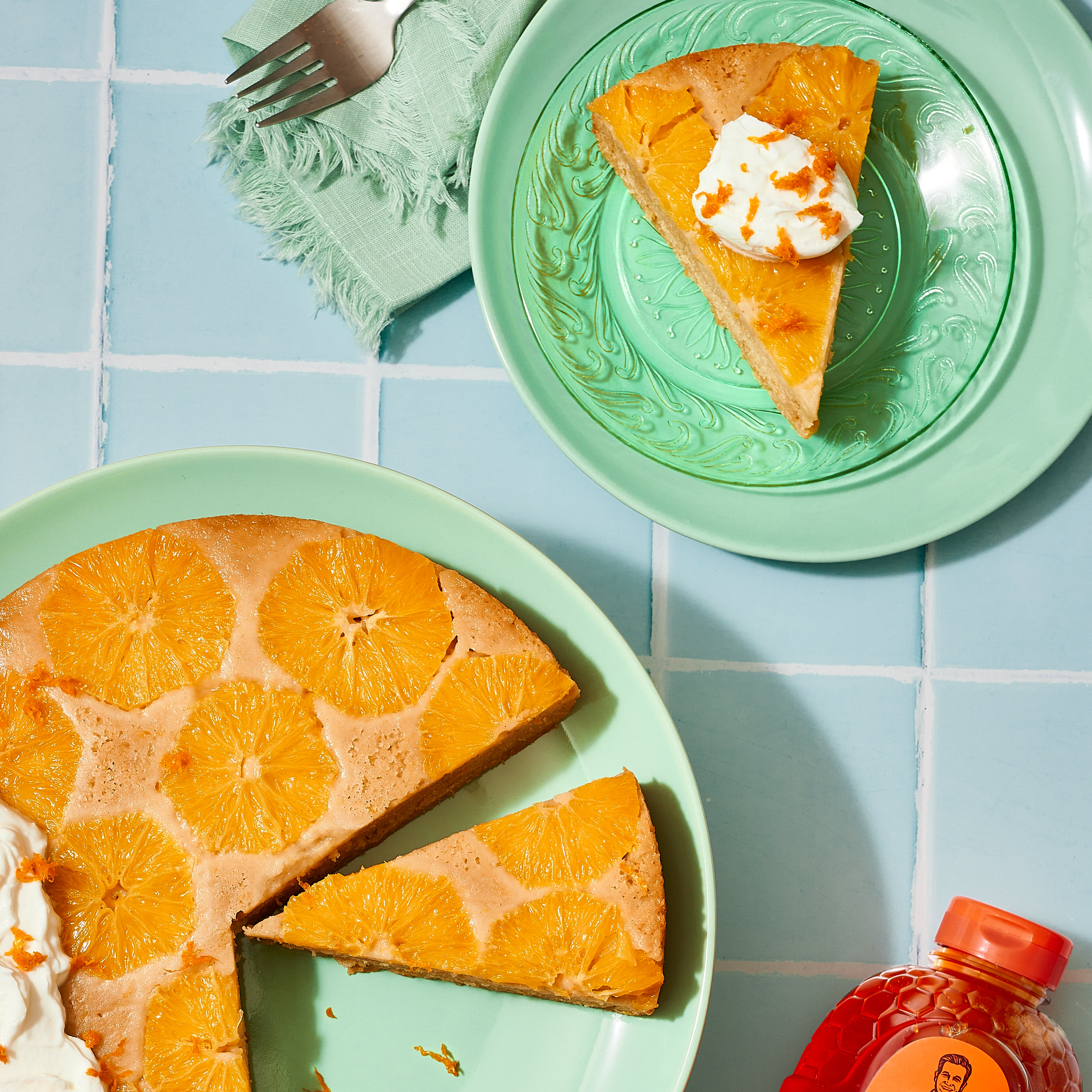 Olive oil cake sliced and garnished with cream next to a bottle of Nate’s honey on a pink table.