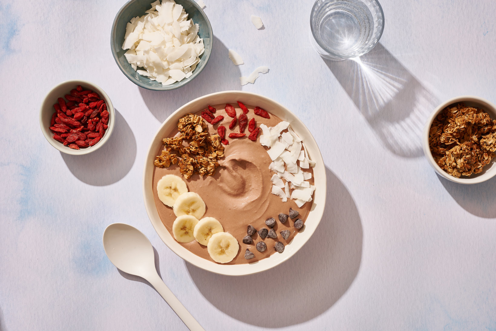 A bowl of chocolate smoothie topped with banana, granola, coconut,  and goji berries with ingredients in small bowls.