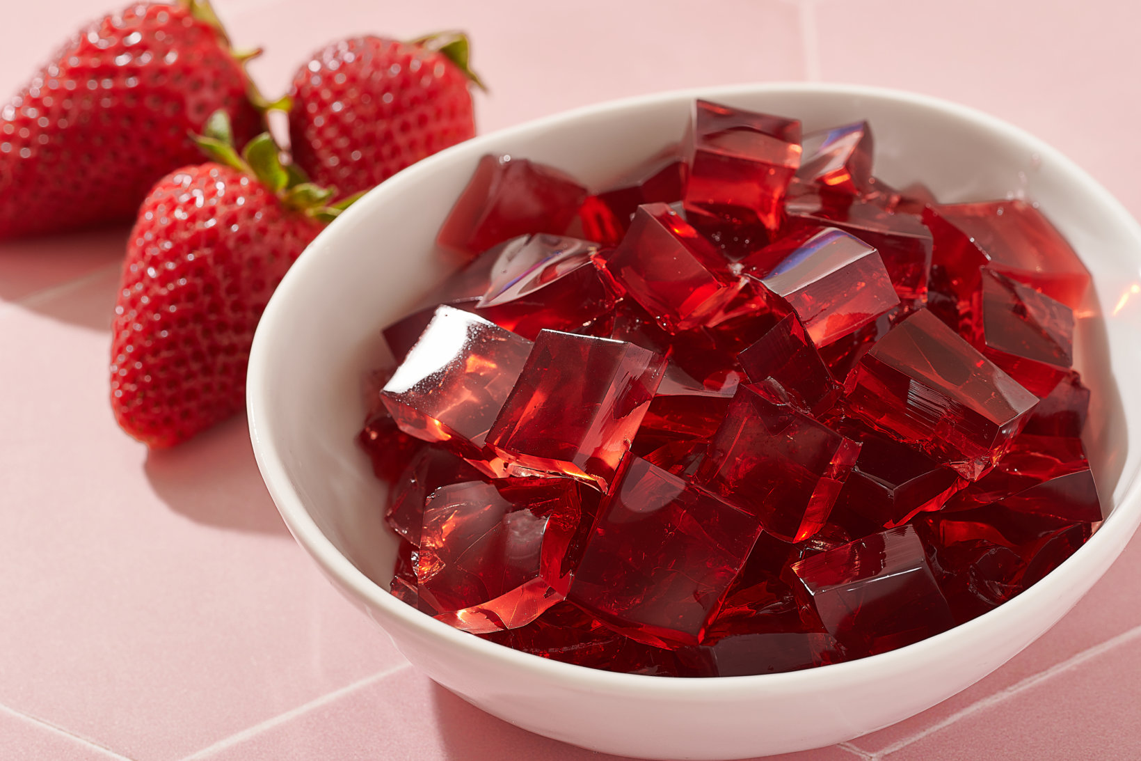 White bowl filled with red gelatin cubes and three strawberries on pink tiled surface.