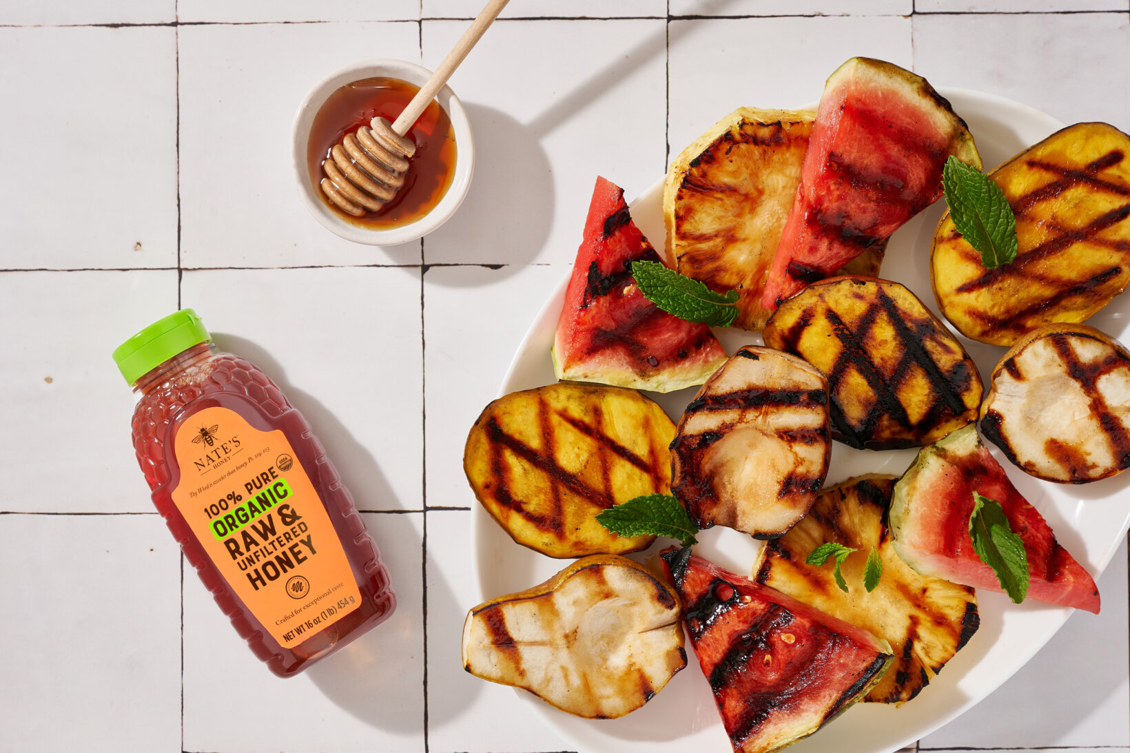 Grilled fruit slices on a plate with mint leaves, a honey dipper and a bottle of Nate’s Organic Honey.
