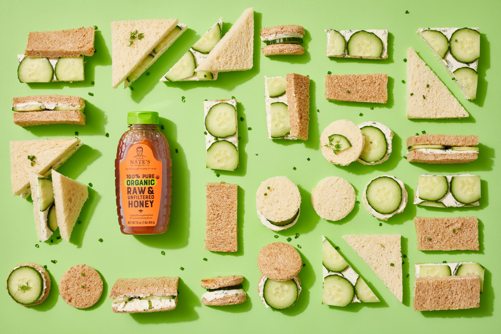 Cucumber sandwiches arranged around a bottle of Nate’s organic honey on a green background.