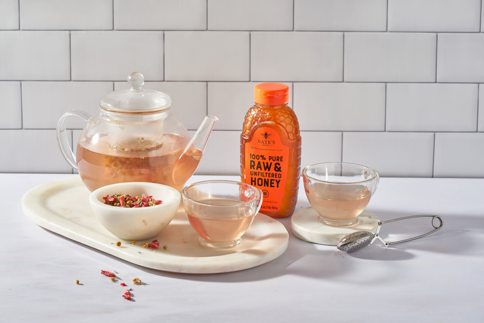 Clear teapot and cups filled with Chamomile tea, with a Nate’s Honey bottle on a white table and subway tile background.
