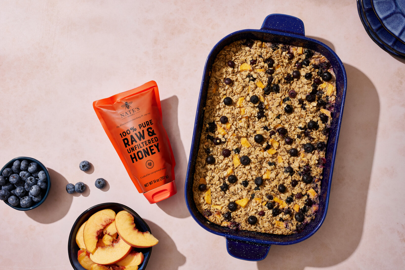 A baked oatmeal dish in a blue dish, a tube of honey, with bowls of blueberries and peach slices on a beige surface.