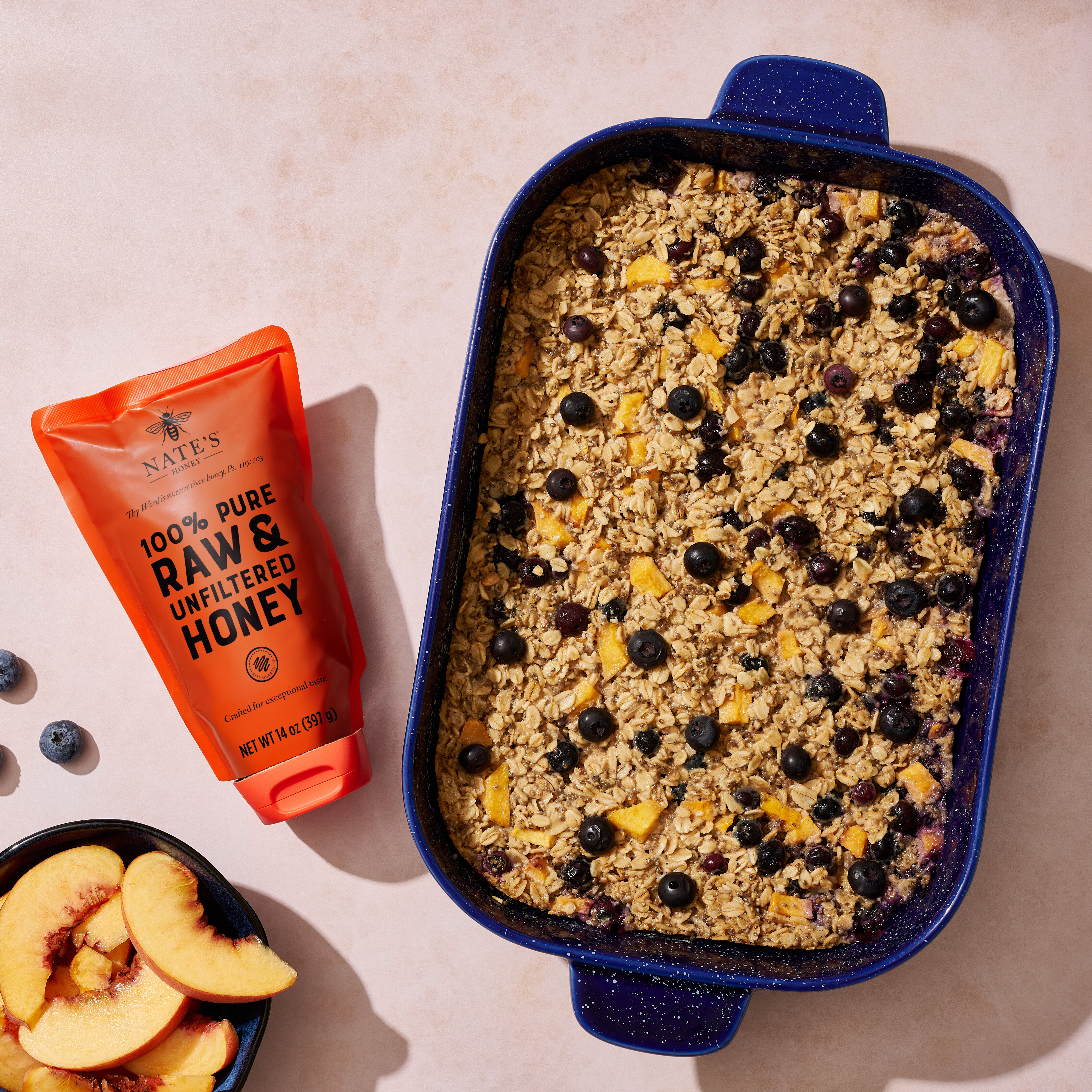 A baked oatmeal dish in a blue dish, a tube of honey, with bowls of blueberries and peach slices on a beige surface.
