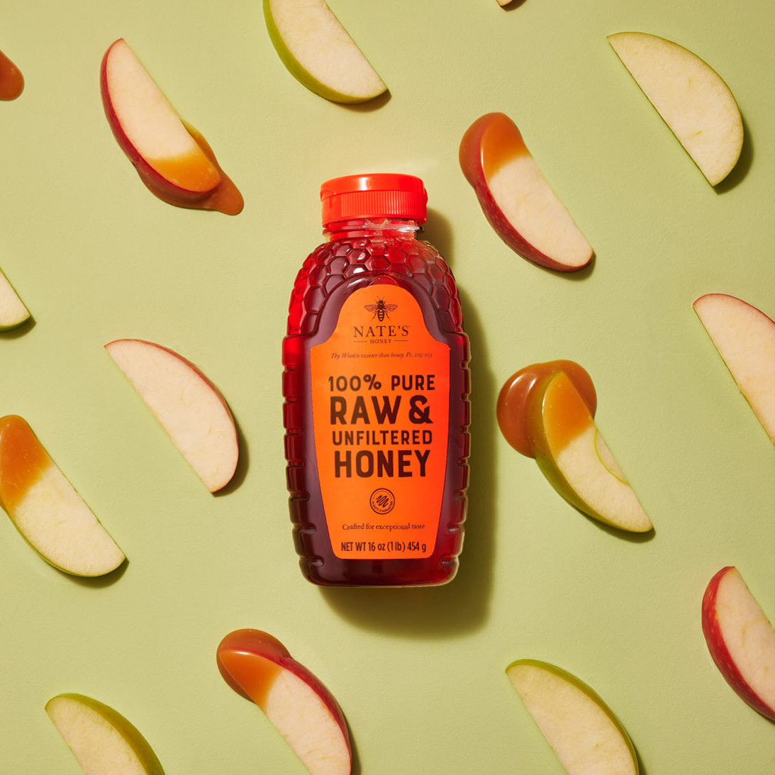 A bottle of Nate's honey with an orange cap surrounded by sliced apple pieces dipped in honey on a green background.