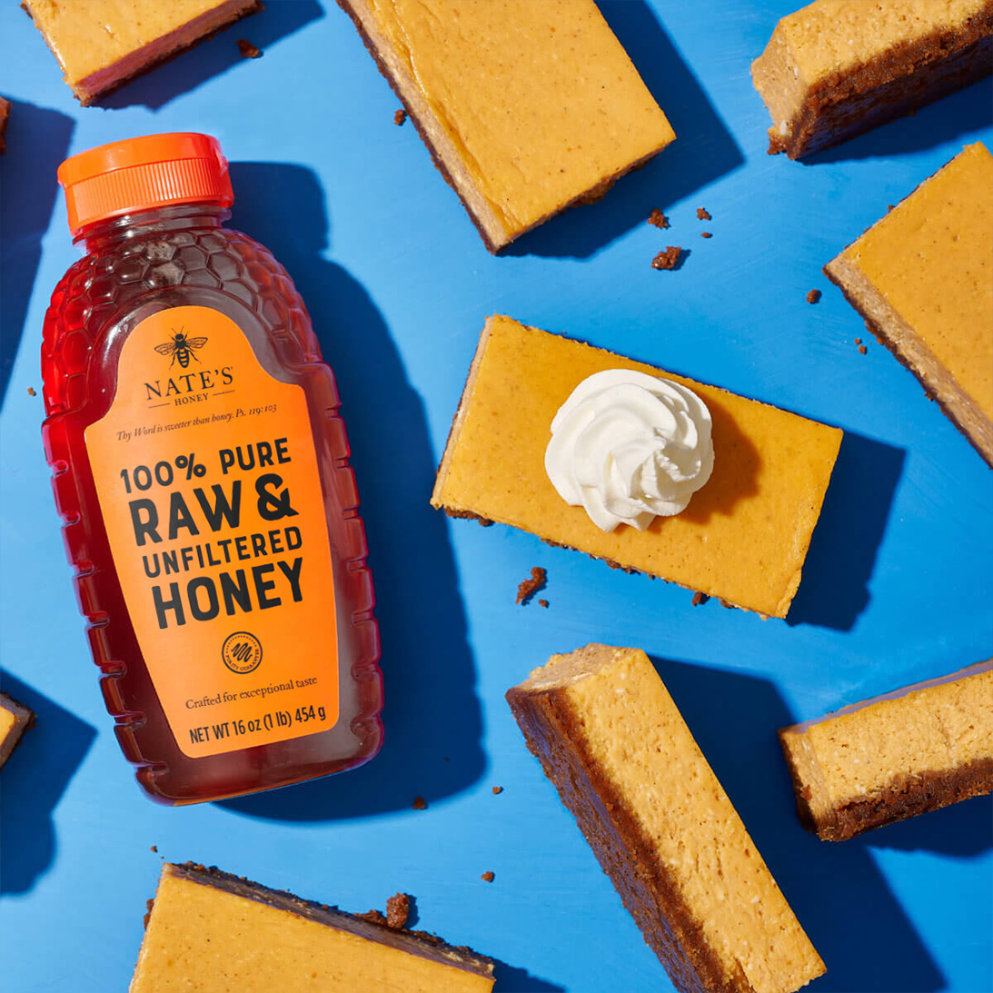 Array of pumpkin bars, some with whipped topping, arranged next to Nate's honey bottle on blue background.
