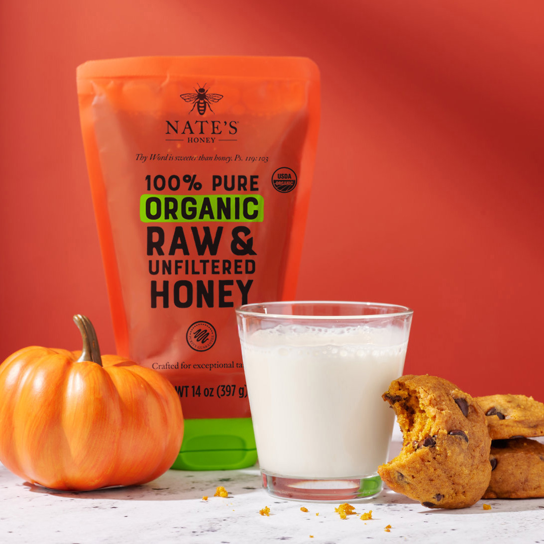 A festive setting with a Nate's organic honey pouch, a glass of milk and pumpkin cookies with a warm backdrop.