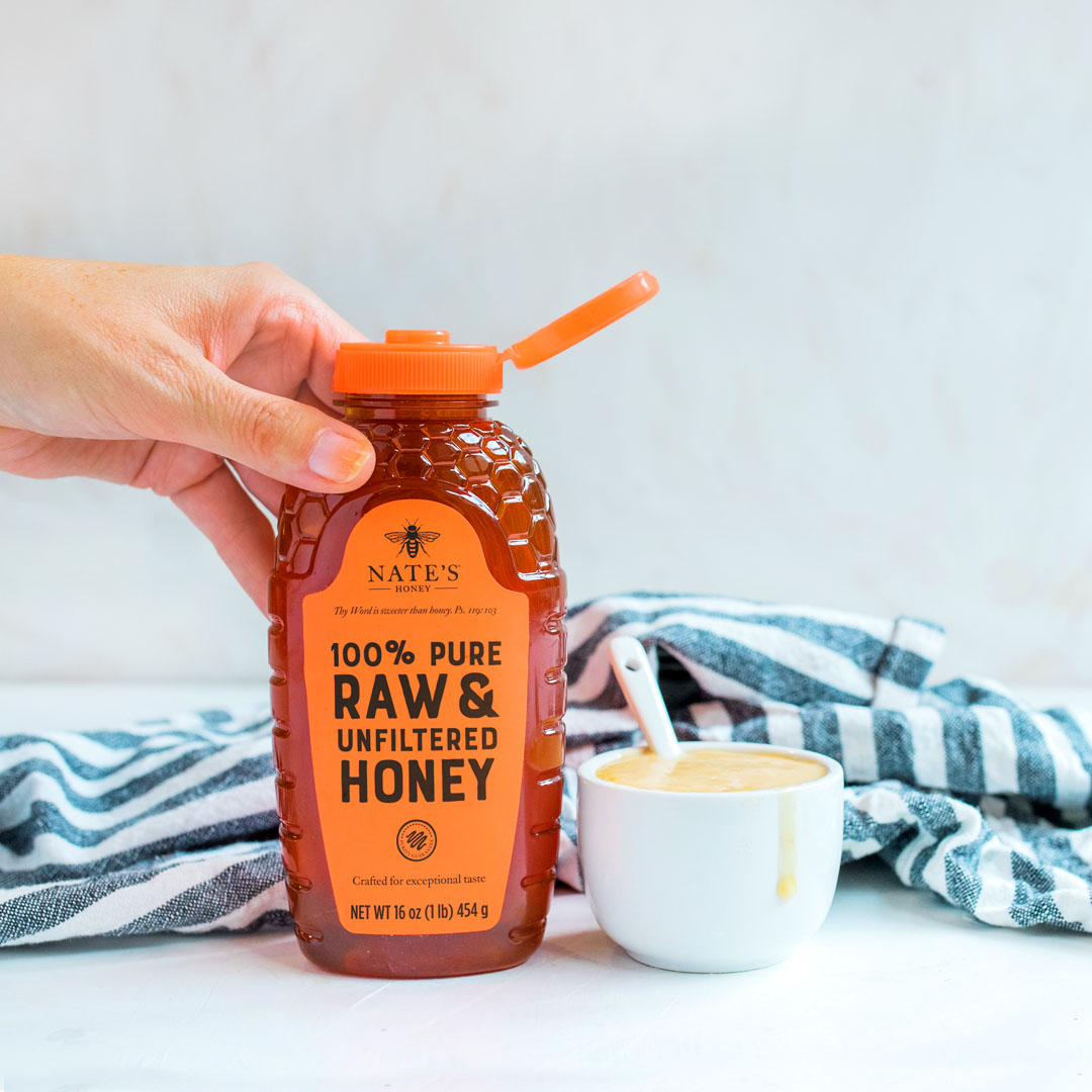 Hand holding a Nate’s Honey bottle, cup with honey mustard and spoon with  striped napkin in background.
