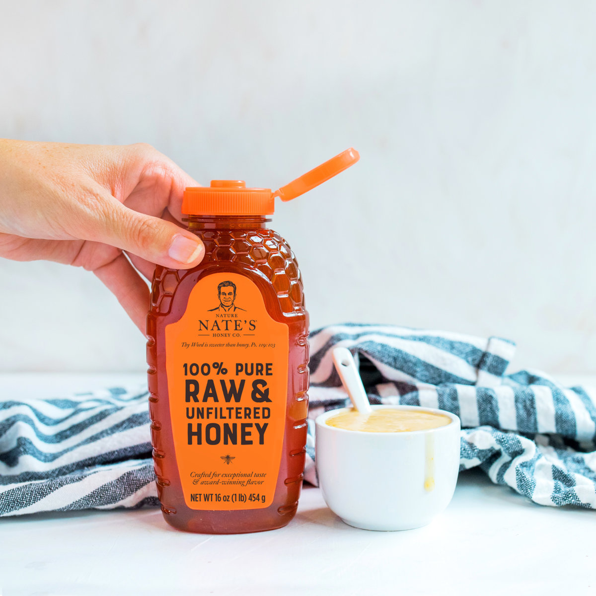 Hand holding a Nate’s honey bottle, cup with honey mustard and spoon with  striped napkin in background.