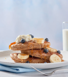 Pancakes with berries on plates, coffee cups, and Nate's honey bottle on a table. plate with pancakes topped with fruit, honey and whipped cream with blue kitchen background