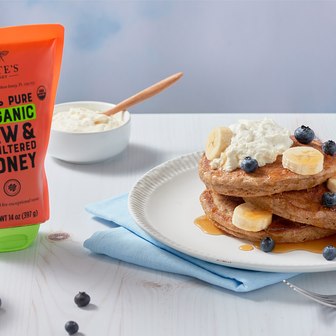 Pancakes with blueberries, bananas and cream beside a Nate’s Organic Honey pouch, blueberries and a glass of milk.