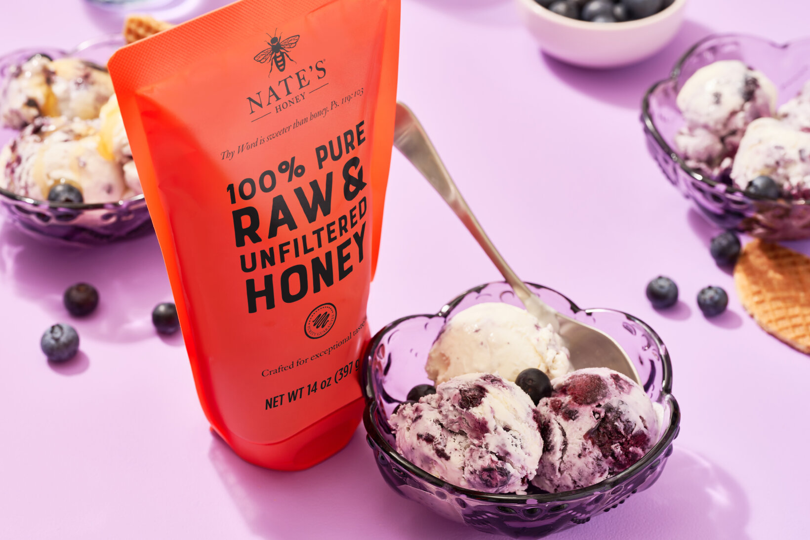 A pouch of Nate's 100% pure raw & unfiltered honey beside a bowl of blueberry ice cream on a purple background.
