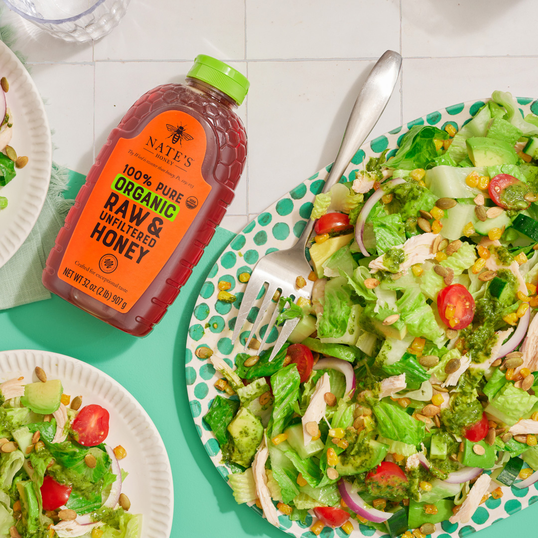 Fresh garden salads with avocado and cherry tomatoes, served next to a bottle of organic Nate’s Honey with a green cap.