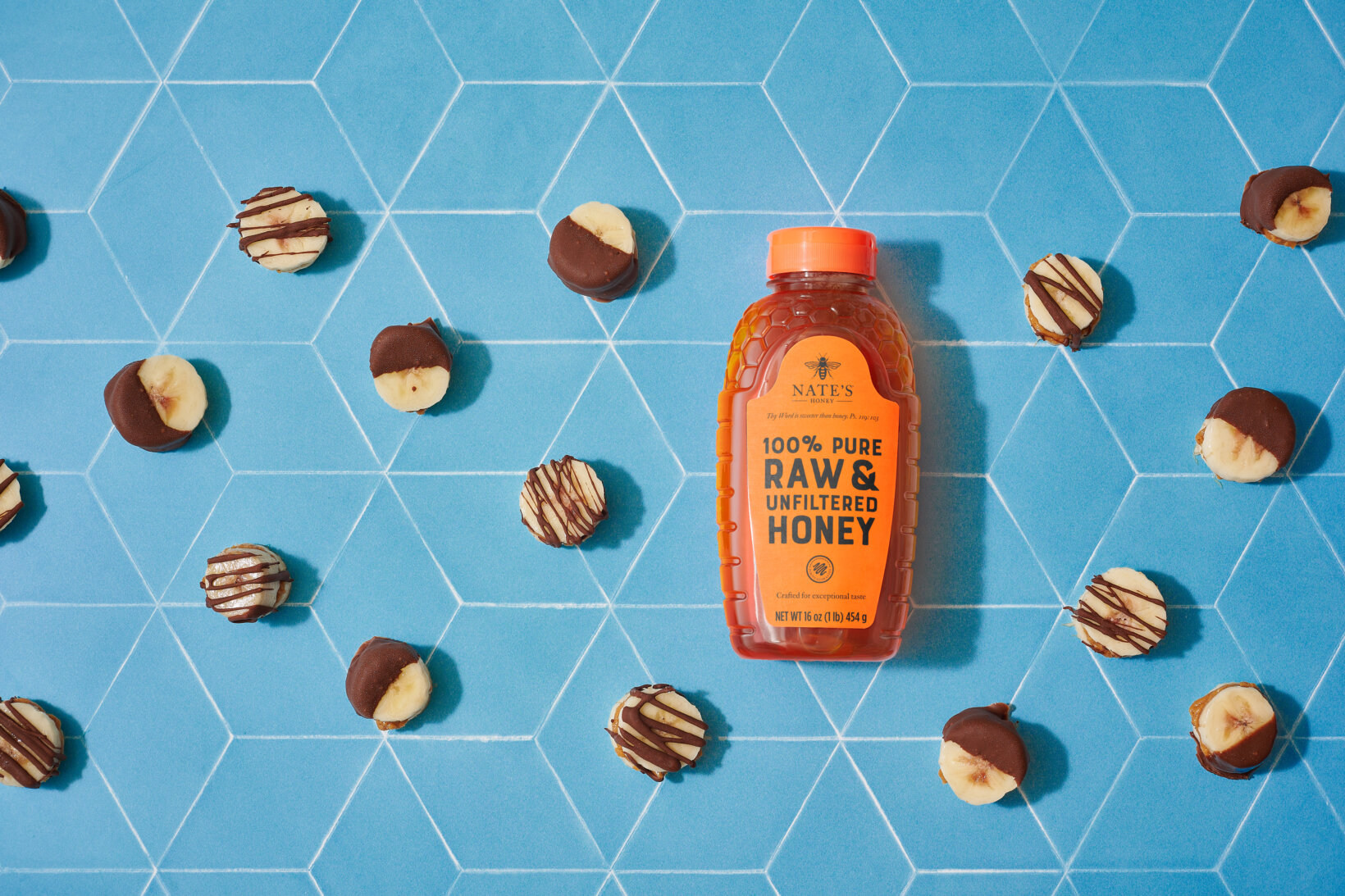 Nate's honey bottle surrounded by chocolate-drizzled banana slices on a blue tile background.