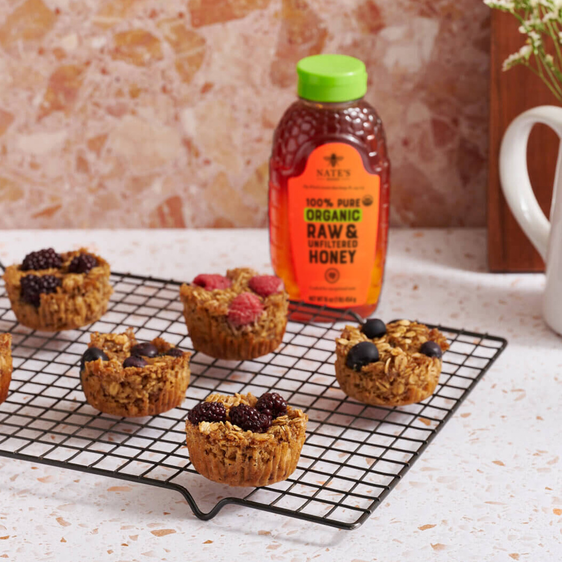 Baked oatmeal cups with berries on a wire rack next to a bottle of Nate’s organic honey and a pitcher of flowers.