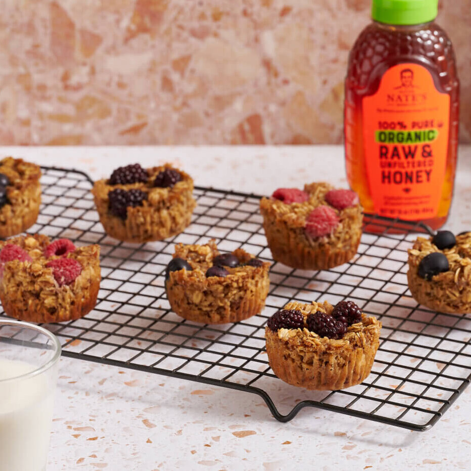 Baked oatmeal cups with berries on a wire rack next to a bottle of Nate’s organic honey and a pitcher of flowers.