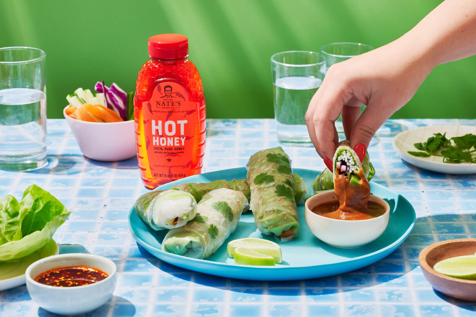 A hand dipping a spring roll in sauce with a bottle of Nate’s hot honey and fresh veggies on a blue patterned table.