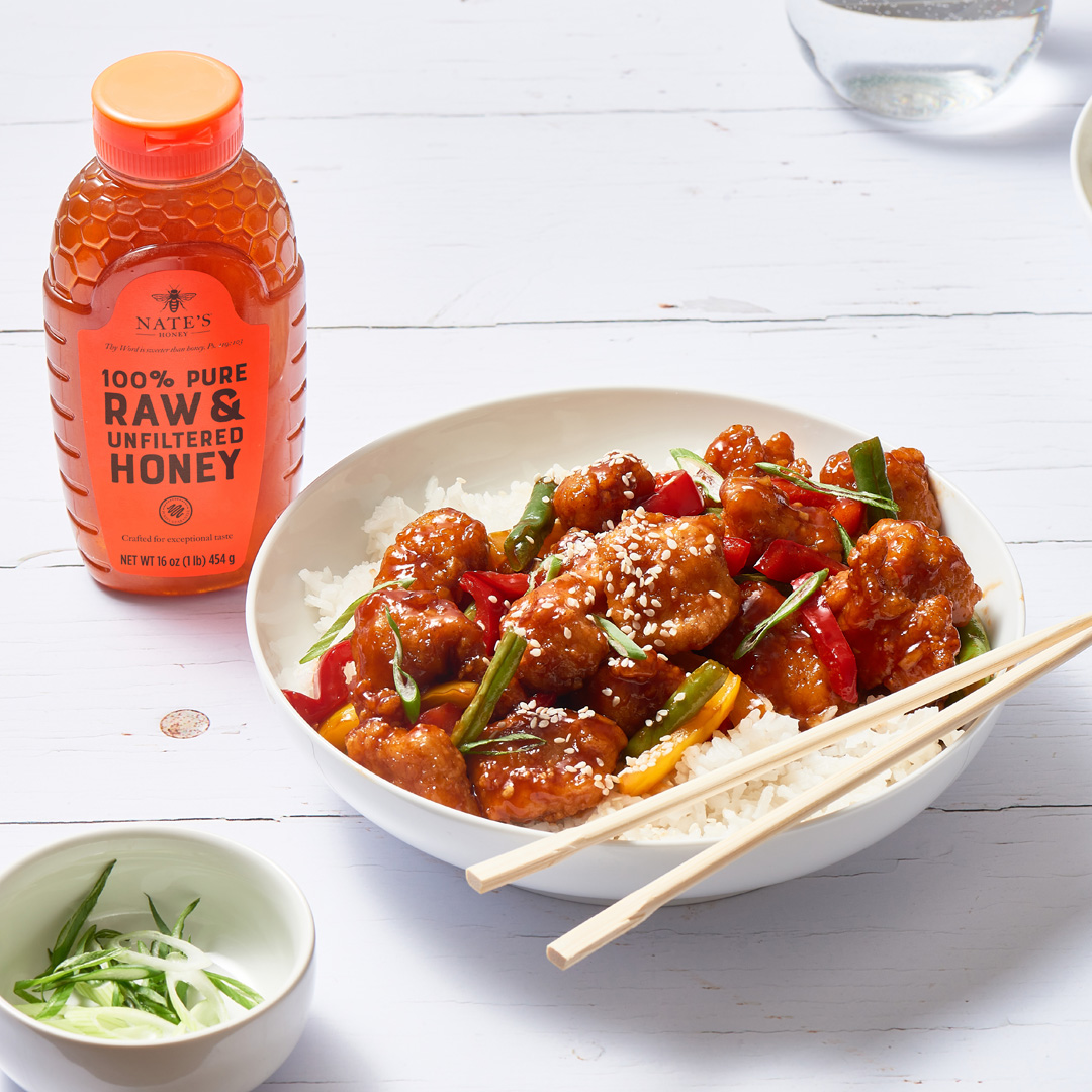 A bottle of Nate’s Honey next to a plate of sesame chicken with vegetables over rice, suggesting a honey-infused recipe.