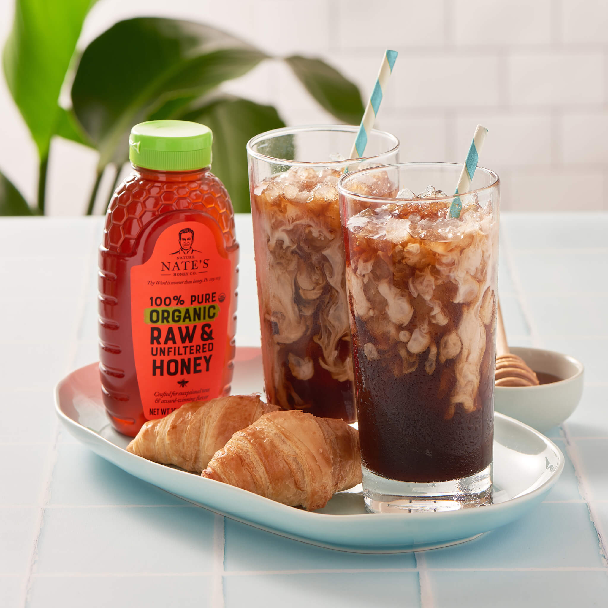 Two glasses of iced coffee with straws, a bottle of Nate’s organic honey and two croissants on a tray.