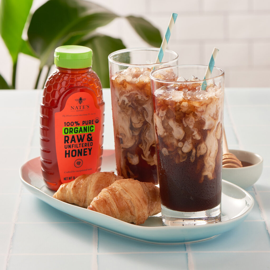 Iced coffee with mint, cream pitcher, and a honey bottle on a bright table. Iced coffees with straws, croissants and a Nate’s Honey organic bottle on a blue-tiled table with a plant backdrop.