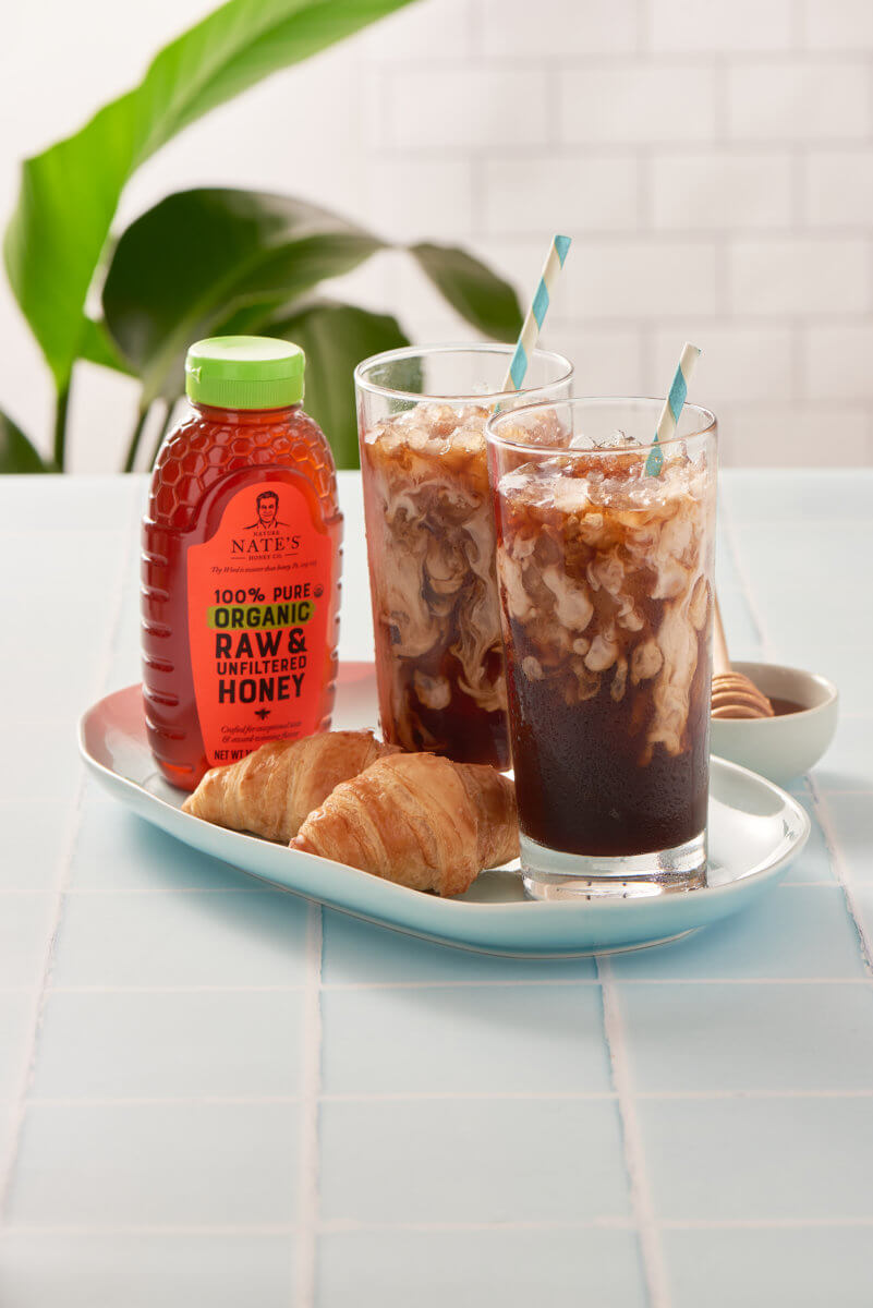 Iced coffees with straws, croissants and a Nate’s organic honey on a blue-tiled table with a plant backdrop.