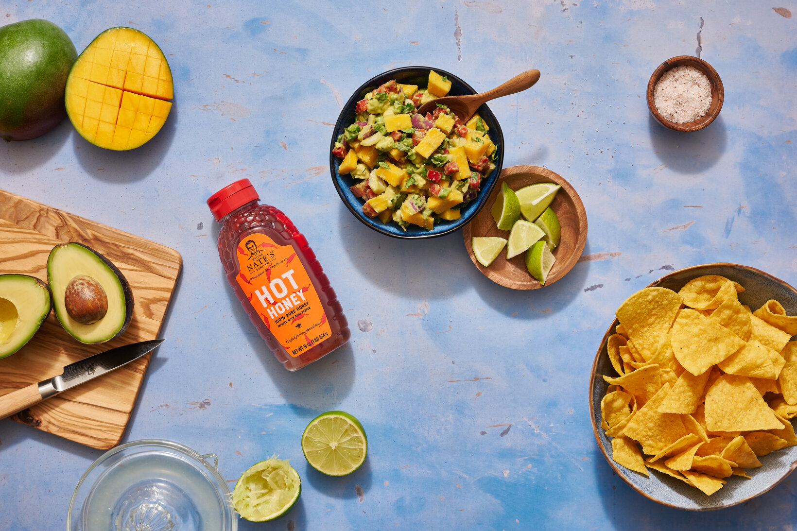 Mango salsa, chips and a bottle of Nate’s hot honey on a blue-speckled background surrounded by fresh ingredients.