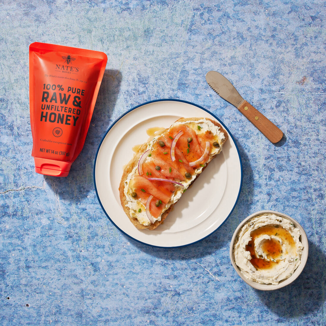 Smoked salmon and cream cheese toast drizzled with honey, next to Nate's honey pouch, knife and bowl on a blue surface.
