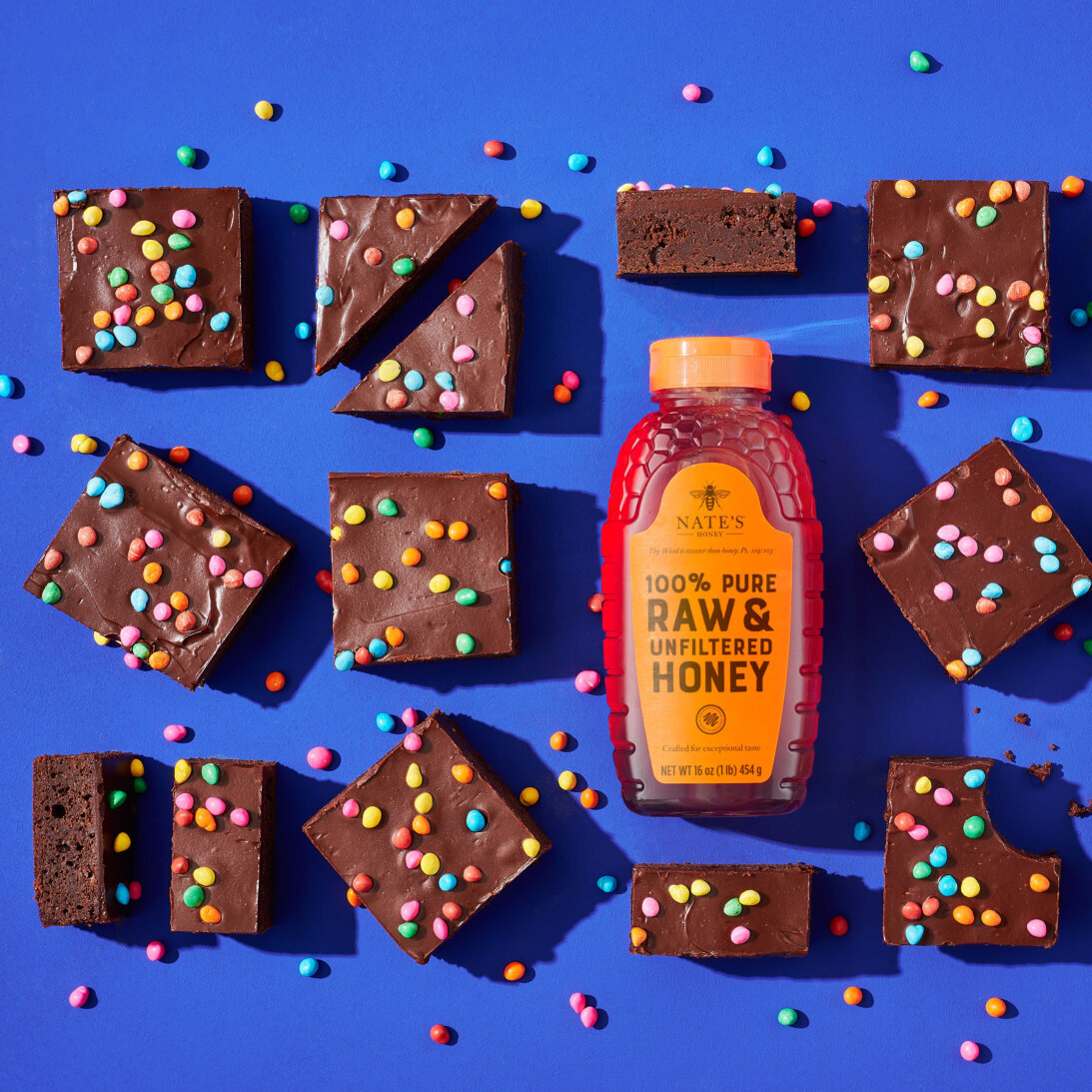 Chocolate brownies with colorful sprinkles and a Nate’s honey bottle on blue background.