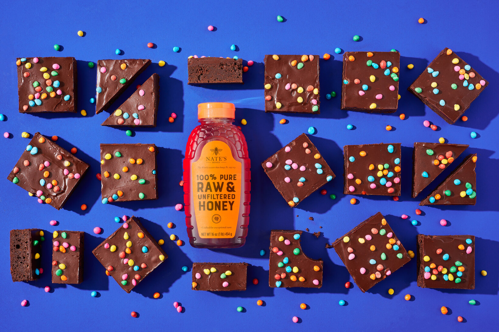 Chocolate brownies with colorful sprinkles and a Nate’s honey bottle on blue background.