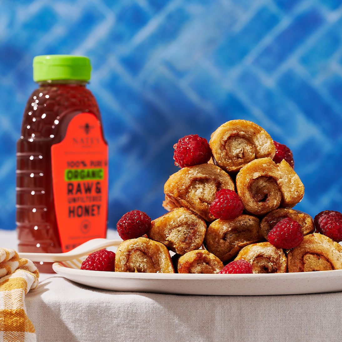 French toast roll-ups with raspberries and a bottle of Nate's organic honey on a white tablecloth.