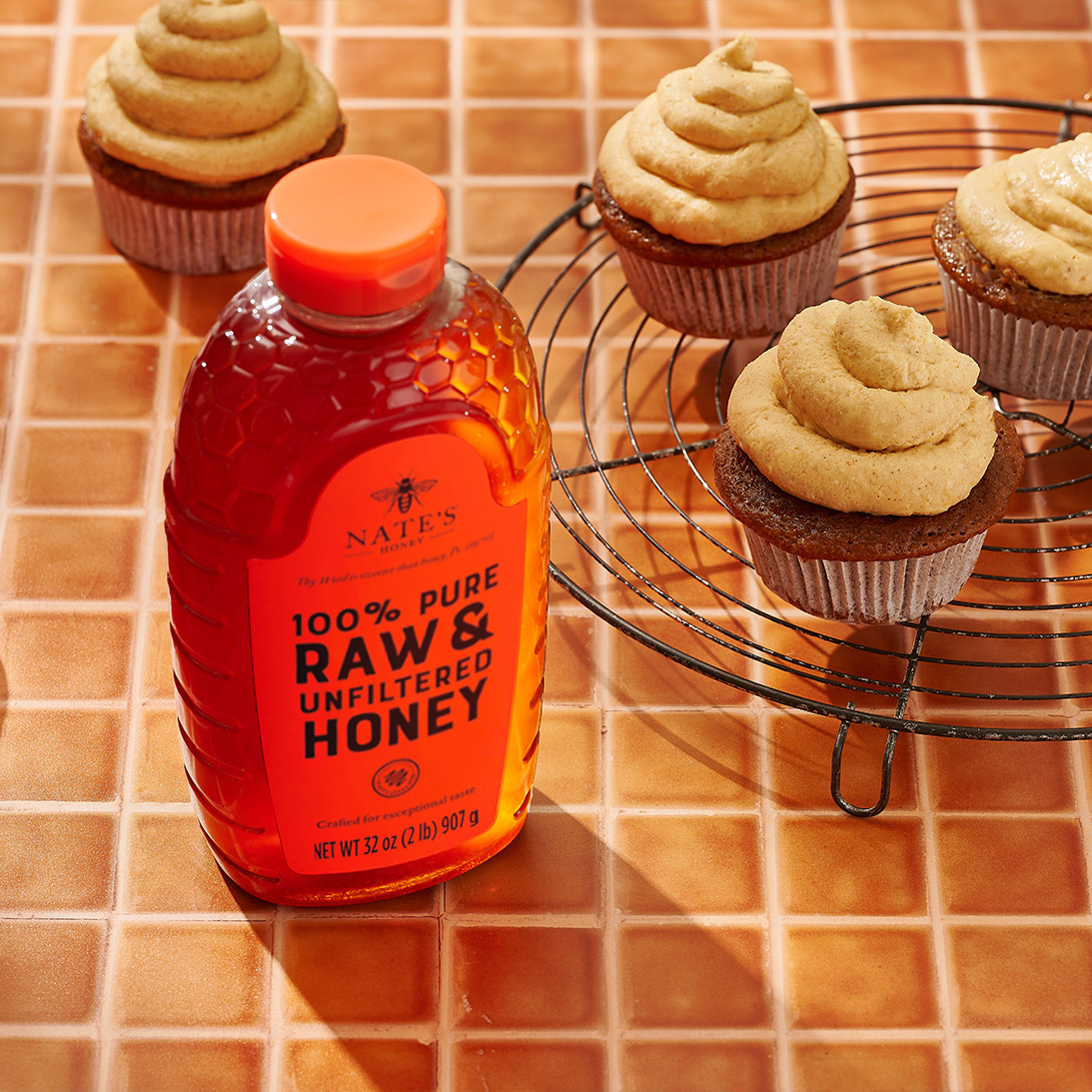Pumpkin cupcakes on a cooling rack and a bottle of Nate’s raw & unfiltered honey on a tiled orange surface.